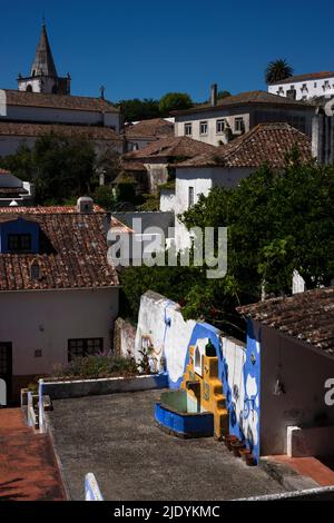 Farbenprächtiger Brunnen im Vordergrund mit Blick auf die pantiled Dächern und ummauerten Gärten im historischen Zentrum der mittelalterlichen ummauerten Stadt Óbidos, Centro, Portugal, Blick auf den hockenden Turm der Igreja de Santa Maria, die Kirche aus dem 15.. Jahrhundert auf dem Hauptplatz, die Praça di Santa Maria. Stockfoto