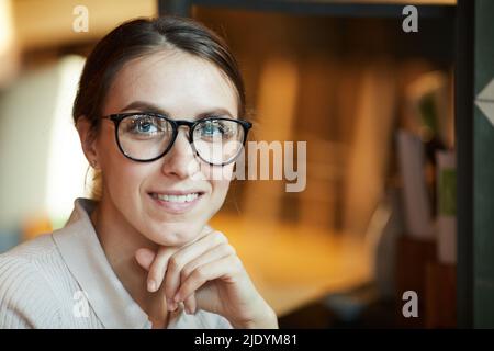Positive junge hübsche Dame mit freundischem Lächeln, die eine stilvolle Brille trägt, die den Kopf an die Hand lehnt und die Kamera anschaut Stockfoto