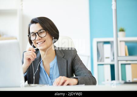 Fröhliche, aufgeregte junge Geschäftsfrau in einer Brille, die das Mikrofon der Kopfhörer benutzt, während sie mit dem Geschäftspartner via Video-Link interagiert Stockfoto
