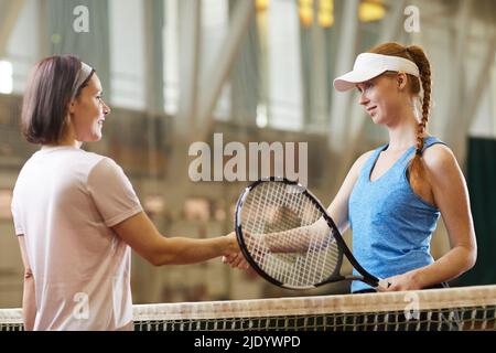 Positive Tennisspielerinnen mittleren Alters in Sportkleidung, die am Netz stehen und sich die Hände schütteln, während sie sich vor dem Spiel gegenseitig begrüßen Stockfoto