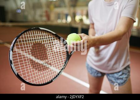 Nahaufnahme einer nicht erkennbaren Frau in einem losen T-Shirt, die auf dem Hallenplatz steht und grünen Ball hält, während sie sich auf den Tennisball vorbereitet Stockfoto