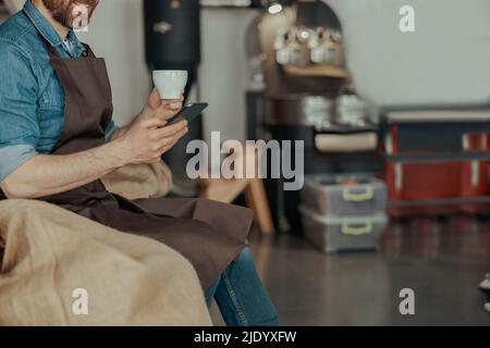 Nahaufnahme des Barista mit einer Tasse Kaffee und Telefon sitzen im Lager der Kaffeefabrik Stockfoto