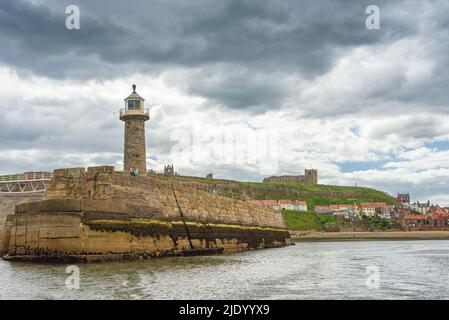 Ein Leuchtturm erhebt sich über einem Pier und hinter einer Kirche steht auf der Skyline. Häuser befinden sich unterhalb der Klippe und ein wolkiger Himmel ist oben. Stockfoto