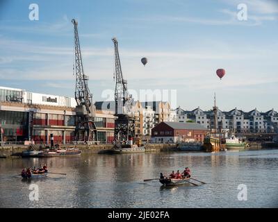 An einem sonnigen Sommerabend schweben Heißluftballons über Kraniche, Ruderboote und den mShed im schwimmenden Hafengebiet von Bristol. Stockfoto
