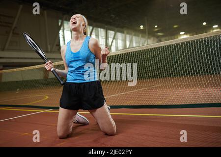 Emotional aufgeregte junge Frau in schweißtreibendem T-Shirt, die auf den Knien steht und schreit, während sie im Tennis Erfolge erzielt Stockfoto