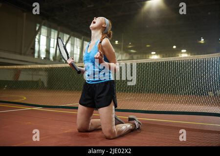 Frustrierte junge Rotschopf-Frau mit Tennisschläger, der auf den Knien steht und weint, während sie das Tennisspiel verliert Stockfoto