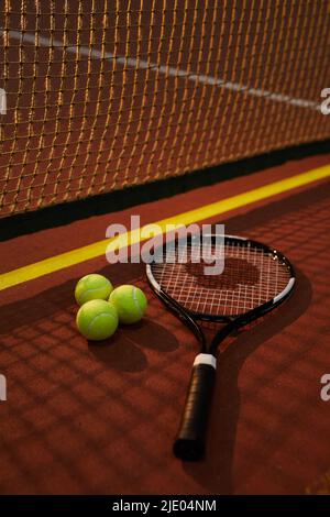 Nahaufnahme eines gut saitieren Spannungsrackes und drei gelben Kugeln, die auf dem dunklen Tennisplatz liegen, Schatten des Netzeffekts Stockfoto