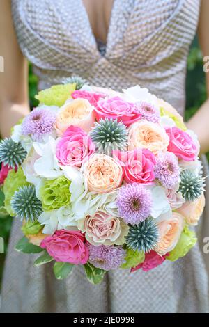 Junge Frau in einem schönen Elfenbeinkleid und mit einem eleganten Brautstrauß in verschiedenen Farben, Rosen, rosa Chrysanthemen und blauer Distel Stockfoto