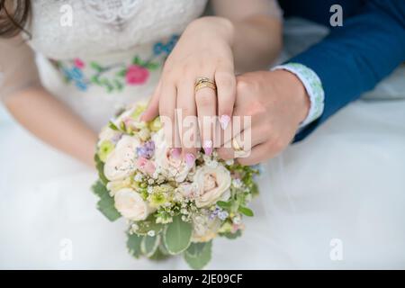 Braut in einem eleganten weißen Kleid und Bräutigam in blauem Anzug mit den Händen über einem zarten Blumenarrangement, das Verlobungs- und Hochzeitsbänder zeigt Stockfoto