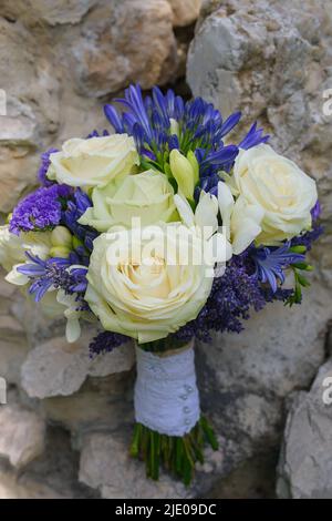 Schöner Hochzeitsstrauß mit elfenbeinweißen Rosen, Elfenbeinfreesien und eleganten blauen afrikanischen Lilien, die vor einem felsigen Hintergrund ruhen Stockfoto