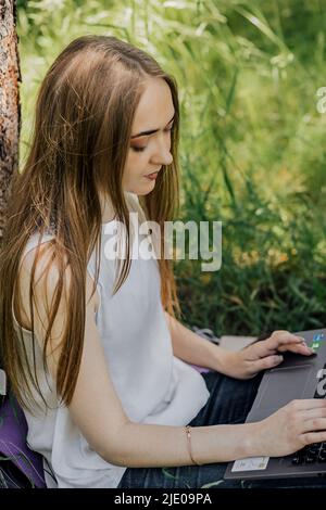 Das Konzept der Fernarbeit. Ein junges Mädchen arbeitet mit einem Laptop an der frischen Luft im Park und sitzt auf dem Rasen. Arbeiten Sie als Freiberufler. Das Mädchen nimmt c Stockfoto