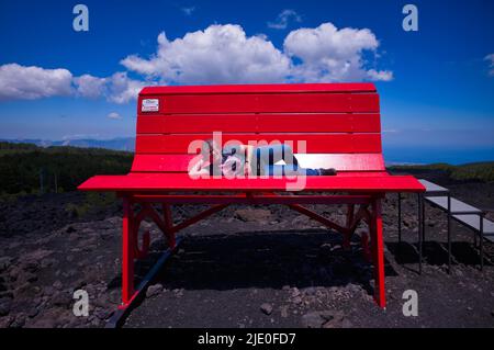 Ältere Frau auf riesiger roter Bank liegend, Big Bench Community Project, Big Bench Number 200, Etna, Grande Panchina, Linguaglossa, Sizilien, Italien Stockfoto