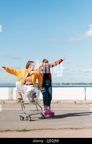 Junge Frau, die im Einkaufswagen in der Nähe von Freunden sitzt, die im Freien Rollschuhe reiten Stockfoto