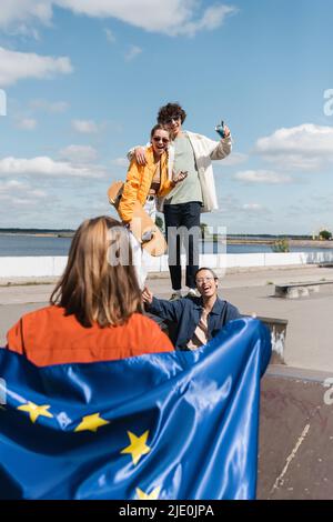 Fröhliche multikulturelle Freunde, die auf eine Frau mit der Flagge der europäischen Union zeigen, die im Vordergrund unscharf ist Stockfoto