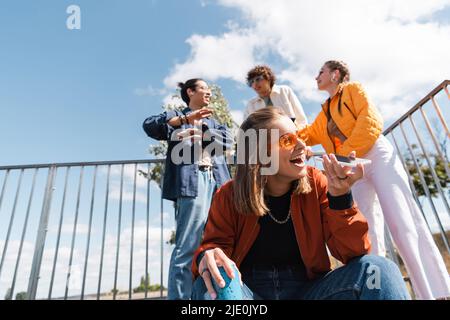 Aufgeregte Frau in Sonnenbrillen, die eine Sprachnachricht in der Nähe multiethnischer Freunde auf verschwommenem Hintergrund sendet Stockfoto