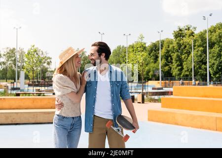 Seitenansicht einer lächelnden Frau, die ihren jungen Freund mit Longboard im Skatepark umarmt Stockfoto