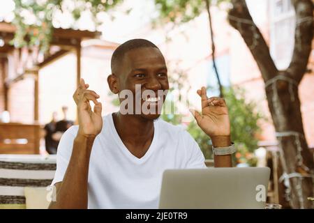 Lächelnder afroamerikanischer Geschäftsmann arbeitet im Café am Laptop, kreuzt die Finger, wünscht Viel Glück. Abschluss des Vertrags Stockfoto