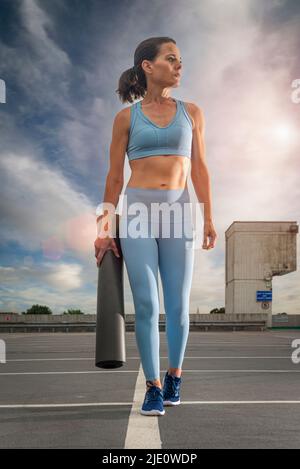 Sportliche Frau, die eine aufgerollte Yoga-Matte trägt, urbaner Hintergrund. Stockfoto