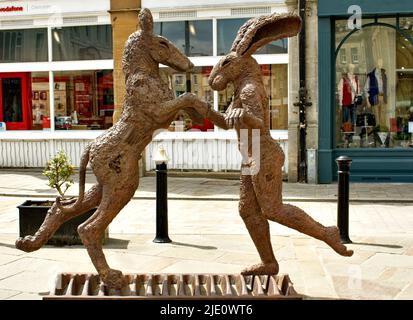 STATUE VON CIRENCESTER GLOUCESTERSHIRE ENGLAND AUF DEM WEST MARKET PLACE MIT EINEM HASENHUND, DER MIT EINEM GROSSEN BRAUNEN HUND TANZTEN Stockfoto