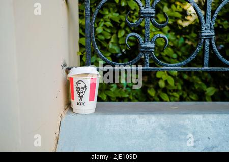 Moskau. Russland. 22. Juni 2022. Leere Kaffeetasse aus Papier mit KFC-Fast-Food-Restaurant-Logo. Ein gebrauchter KFC-Papierbecher auf der Straße in der Nähe eines schmiedeeisernen Zauns. Stockfoto