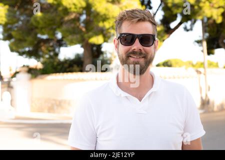 Außenaufnahme eines schönen bärtigen jungen Mannes mit Sonnenbrille, der an warmen sonnigen Tagen über dem Stadtgarten posiert und weißes Poloshirt trägt Stockfoto