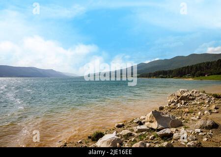 Transparenter türkis tiefer See mit felsigen leeren schmalen Ufer vor dem Hintergrund der hügeligen grünen Bergkette mit dichtem Fichtenwald bedeckt, a c Stockfoto