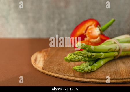 Zweige von frischem grünem Spargel auf einem Holzbrett, roter großer Pfeffer auf dem Hintergrund. Brauner Hintergrund. Grundlegendes Trendkonzept mit Kopierbereich Stockfoto