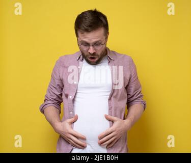 Der Mensch ist von seinem Übergewicht schockiert, er sieht sich das überschüssige Bauchfett an und hält es mit den Händen. Gesundes Lifestyle-Konzept. Stockfoto