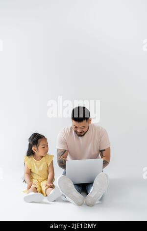 Glücklich asiatischen Vater arbeiten Remote auf Laptop in der Nähe Tochter auf grau Stockfoto
