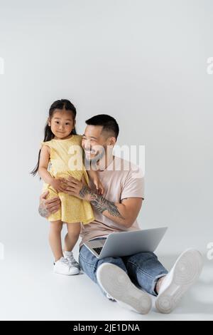 Fröhlicher asiatischer Vater sitzt mit Laptop in der Nähe der Tochter auf grau Stockfoto