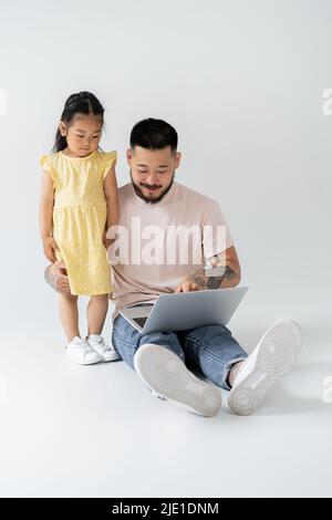Fröhlicher asiatischer Vater mit Laptop in der Nähe Tochter in gelbem Kleid auf grau Stockfoto