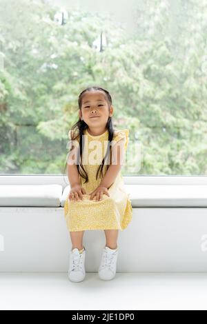 Volle Länge der fröhlichen asiatischen Mädchen in gelbem Kleid sitzt auf weichem Kissen auf Fensterbank Stockfoto