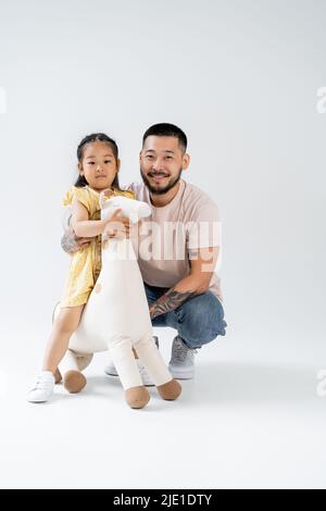 In voller Länge glücklich asiatische Kind hält Spielzeug Giraffe und in der Nähe tätowiert Vater auf grau Stockfoto