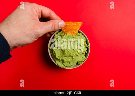 Die Hand des Mannes taucht ein Dreieck aus Mais in Guacamole in eine Keramikschale auf einen runden Schieferteller, roter Hintergrund Stockfoto