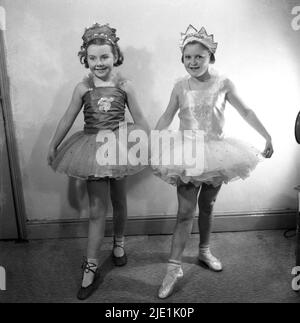 1950s, historisch, zwei kleine Mädchen in ihren Ballettkostümen, spitzenschuhen und Kronen auf ihrem Kopf, die in Innenräumen posieren, Stockport, England, Großbritannien. Das Ballettkostüm, ein Rüschenrock, der an einem schnittig anliegenden Oberteil befestigt ist, wird auch als Tutu bezeichnet. Stockfoto