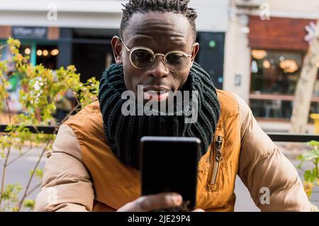 Junger afrikanischer Mann mit Brille, schwarzem Schal und kurzen Haaren, draußen vor der Cafeteria sitzend, lächelnd mit erhobenen Augenbrauen und mit Blick auf das Telefon Stockfoto