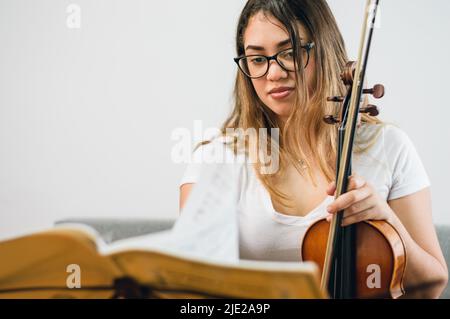 Junge lateinische kaukasische Geigerin, mit Brille ethnisch venezolanischen langen Haaren, zu Hause Studium der Noten sitzen auf dem Sofa mit Geige in der Hand, Polizist Stockfoto