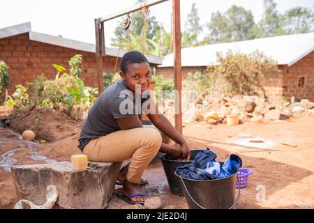 Junge afrikanische Mädchen wäscht Kleidung draußen mit Waschseife und einem Eimer Stockfoto