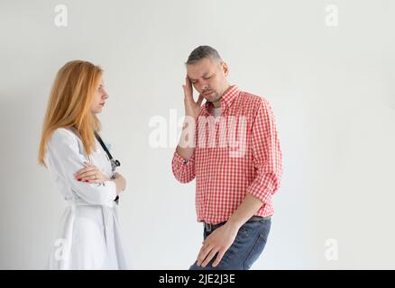 Eine Ärztin konsultiert einen Mann mit starken Kopfschmerzen auf einem hellen isolierten Hintergrund. Männliche Migräne. Schreckliche Kopfschmerzen Stockfoto