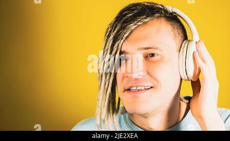 Junger, hübscher Mann mit kabellosen weißen Kopfhörern, der Musik hört. Entspannter Typ mit blonden Dreadlocks, der Lieder hört und auf gelbem Backgroun singt Stockfoto