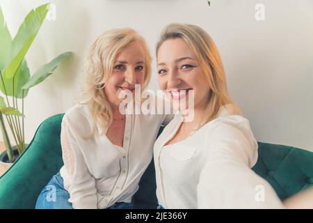 Horizontale Innenaufnahme in einer Wohnung mit zwei blonden, gut gekleideten, attraktiven, tausendweiblichen Geschwistern, die auf einer dunkelgrünen Couch sitzen. Hochwertige Fotos Stockfoto