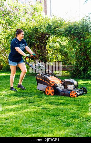 Rückansicht einer Gärtnerin in legerer Kleidung, die gerne frisches Gras mit einem Rasenmäher oder einem Rasenmäher im Hof schneidet Stockfoto