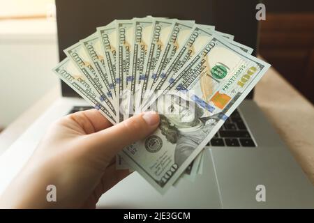 Ein Mann hält einen Fan von hundert Dollar-Scheinen vor dem Hintergrund eines modernen Laptops. Stockfoto