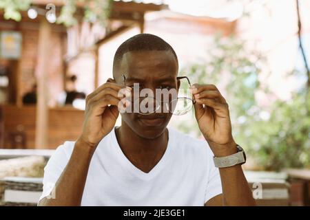 Nahaufnahme müde und müde multirassische, dunkle Haut Mann, der seine Brille auszieht und die Linse überprüft. Sicht- und Sichtprobleme Stockfoto