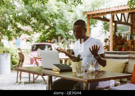 Überrascht und verwirrt afroamerikanischen Mann mit Laptop im Café weiß nicht, was zu tun ist, heben die Hände im Café im Freien Stockfoto