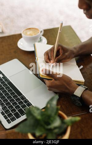 Vertikal zugeschnittenes multirassisches Unternehmen mit Laptop, Notizen im Arbeitsbuch, Kaffee trinken im Straßencafé. Hoher Winkel Stockfoto