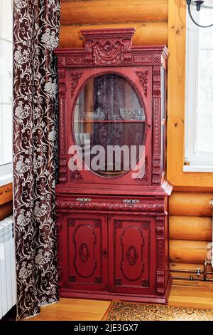 Alter alter, roter Holzschrank mit geschnitzten Dekorationen in der Ecke einer ländlichen Blockhütte zwischen Fenster und braunem Vorhang. Stockfoto