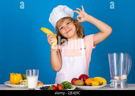 Kinderkoch Koch bereitet Essen halten Banane in isolierten blauen Studio Hintergrund. Kinder kochen. Teen junge mit Schürze und Koch Hut Vorbereitung einer gesunden Stockfoto