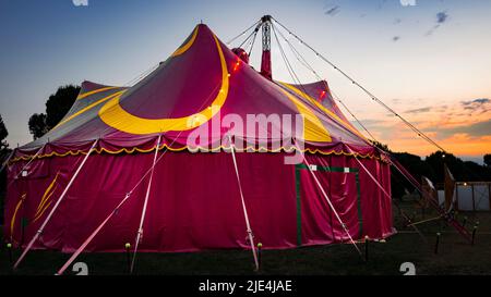 Sonnenaufgang über einem roten, gelben Zirkuszelt, das auf einem Stadtpark steht. Stockfoto