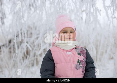 Kaukasisches junges Mädchen mit roten, ruddigen Wangen in warmer Winterkleidung, das die Kamera mit schneebedeckten Bäumen im Hintergrund leicht lächelt. Winterspaziergang Stockfoto